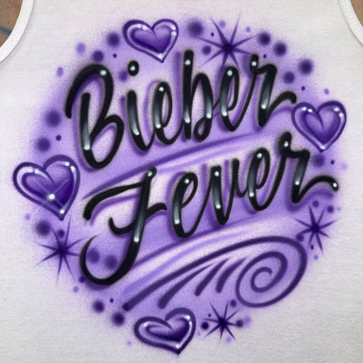 Hand airbrushed Bieber Fever design with purple hearts, swirls, and glowing starburst accents on white apparel.